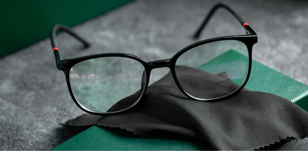 Black rectangular glasses resting on a folded microfibre cloth with a green glasses case on a textured surface.