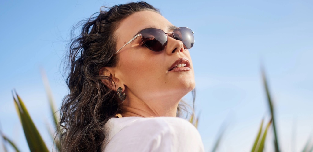 A woman with wavy brown hair wears slim, oval sunglasses with tinted lenses, tilting her face upward toward the sunlight. She has on a white top and decorative earrings, with a clear blue sky and hints of greenery in the background, creating a bright, summery feel.