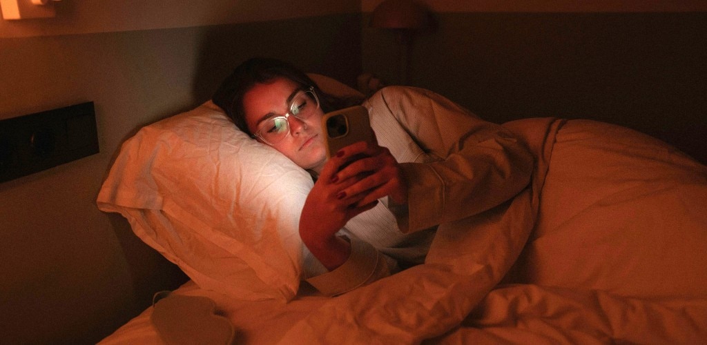 Woman wearing transparent glasses, lying in bed using a smartphone at night, illustrating blue light exposure and its impact on sleep and circadian rhythm.