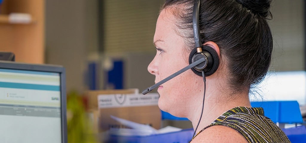 A woman wearing a headset works at a computer in an office setting, viewed from the side as she focuses on the screen.