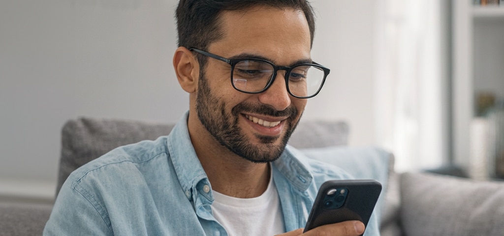 A man wearing glasses smiles while looking at his smartphone, sitting comfortably indoors in a relaxed home setting.