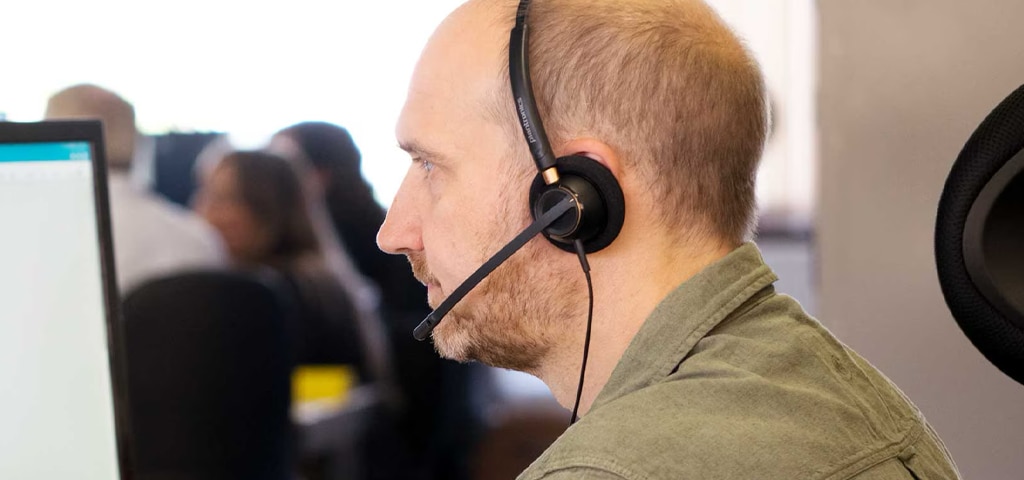 A man with short light hair and stubble wearing a green shirt and headset, sitting at a computer in an office, assisting a customer over the phone.