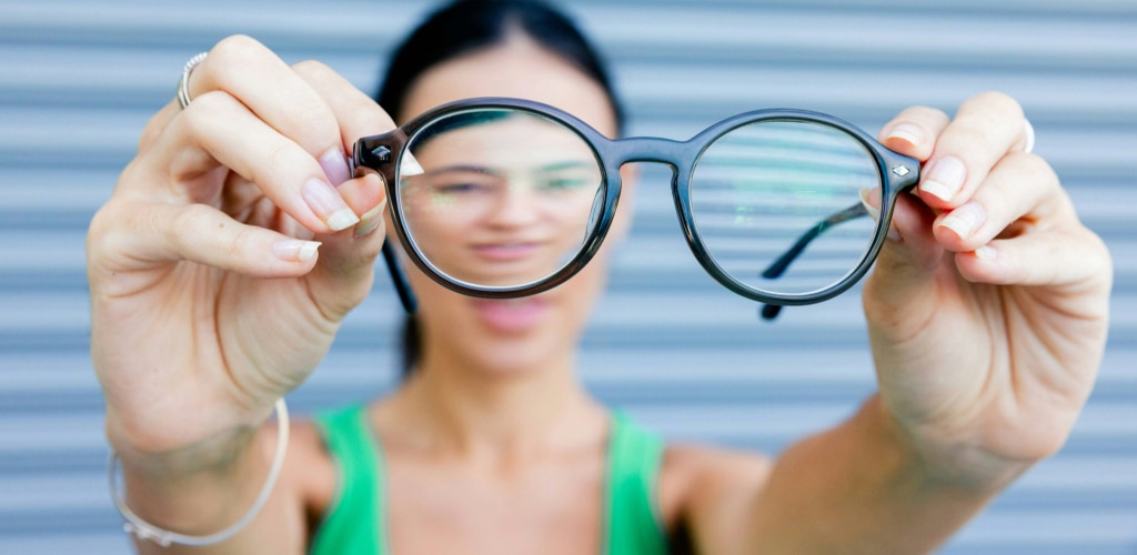 Woman with dark hair holding round black glasses up in front of her face, focusing on the lenses.