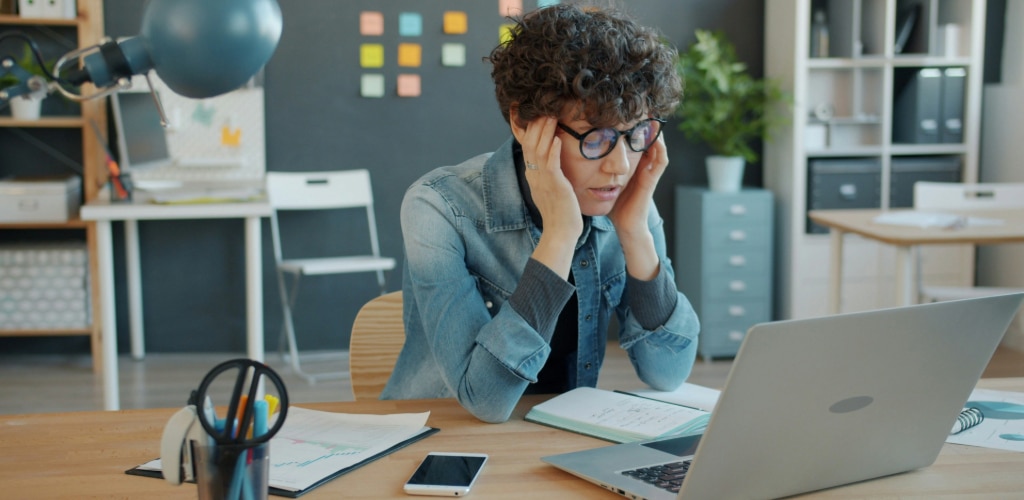 Woman with short curly brown hair wearing round black acetate glasses and a denim shirt over a grey long-sleeve top. She appears to be experiencing digital eye strain while using a laptop, holding her temples and leaning on her desk in a bright, modern office setting. 