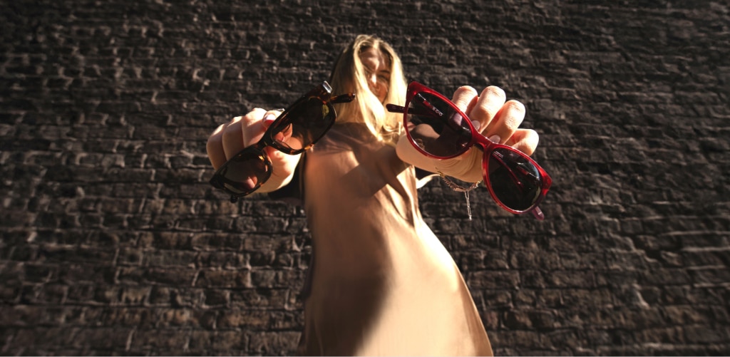 A woman with long blonde hair stands outdoors against a brick wall, holding two pairs of sunglasses toward the camera — one tortoiseshell and one red — creating a bold, close-up perspective with her face slightly out of focus in the background.