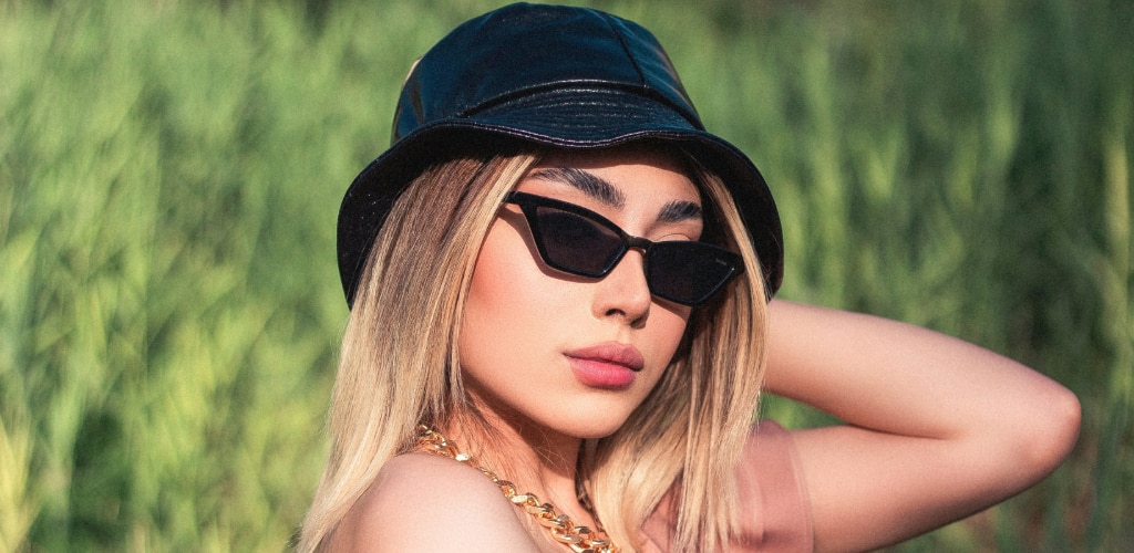 A woman wearing a black leather-style bucket hat and angular cat-eye "it girl" sunglasses poses outdoors against a blurred green background, with one hand raised behind her head and a gold chain necklace visible.
