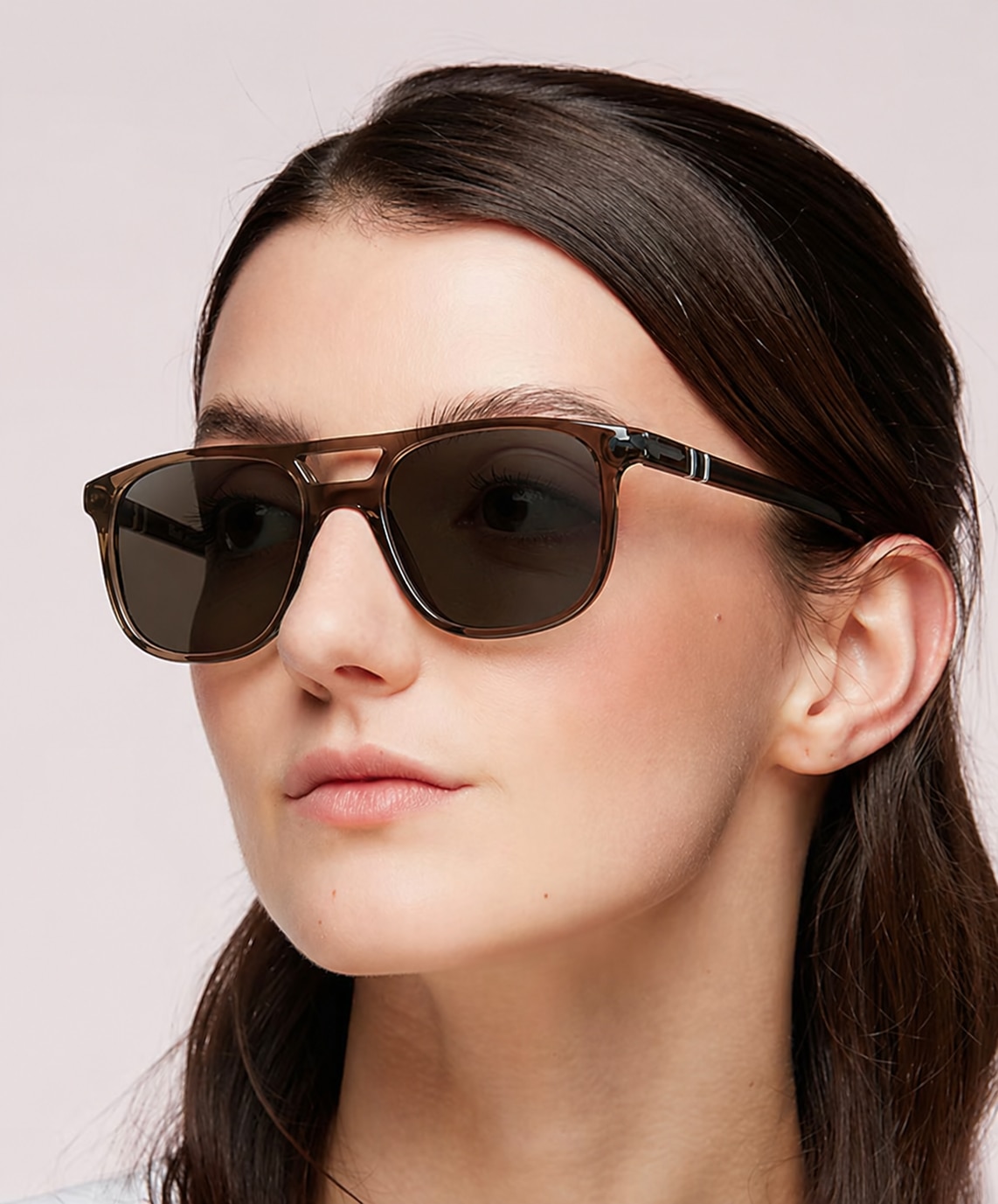 A woman with dark hair wears grey acetate Persol aviator sunglasses, looking slightly to the side against a clean, neutral background.