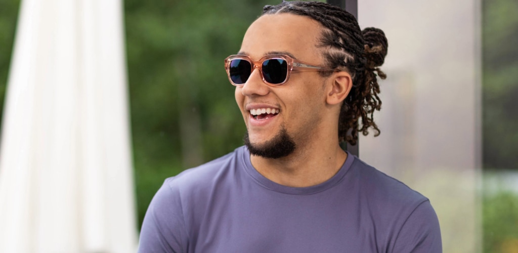 Man with goatee and tied-back hair wearing a blue t-shirt and clear pink glasses with Transitions® lenses, smiling outdoors.