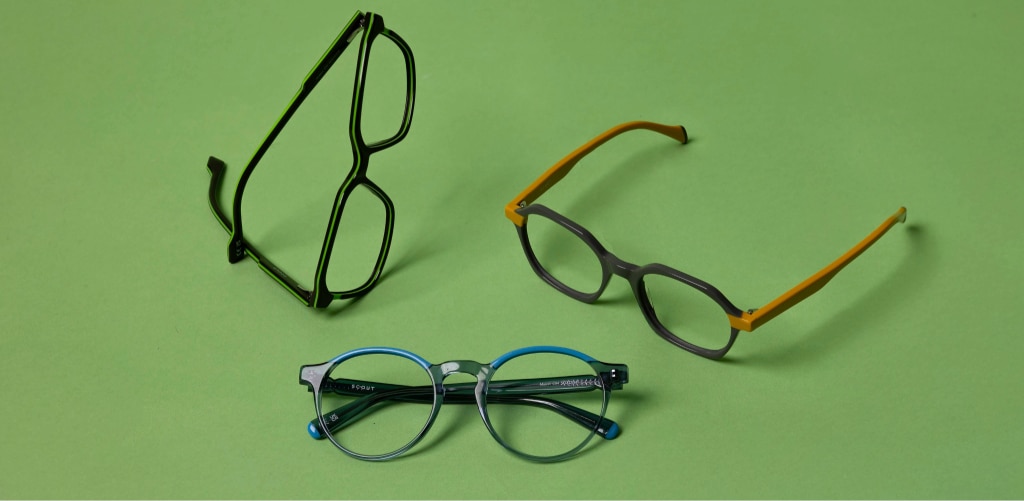 A product shot of three pairs of eyeglasses arranged on a green background. Each frame has a bold, distinctive design: one pair is black with a rectangular shape standing upright, another features a mix of grey and bright yellow accents, and the third is round with blue and transparent tones. The composition highlights variety in colour and style, with soft lighting creating subtle shadows.