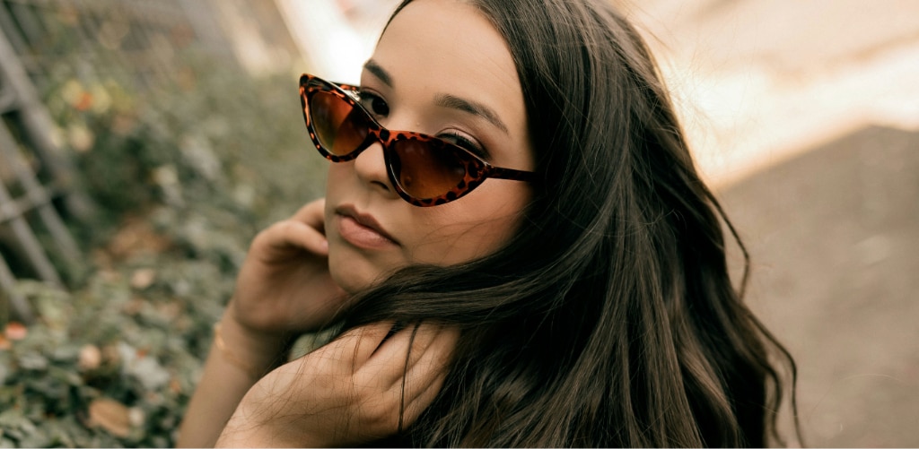 A woman with long dark hair looks over her shoulder while wearing narrow tortoiseshell "it girl" cat-eye sunglasses, with a soft-focus outdoor background of foliage and a path.