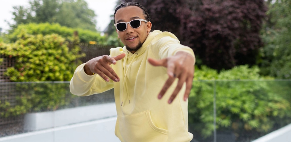 Man with goatee wearing a light yellow hoodie and white-framed Transitions® glasses outdoors, smiling and gesturing, lenses tinted in daylight.