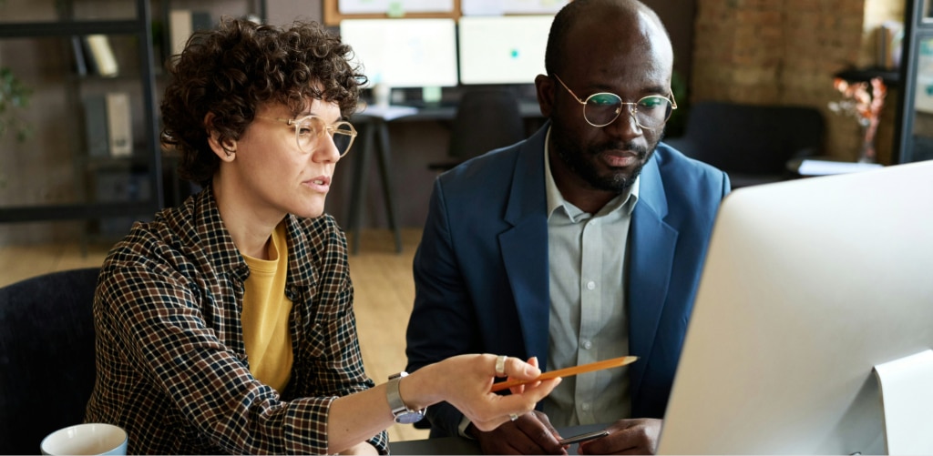 A man in a blue suit and a woman in a check shirt working together at a computer in a softly lit office, illustrating reduced glare and a comfortable screen environment with balanced ambient lighting.