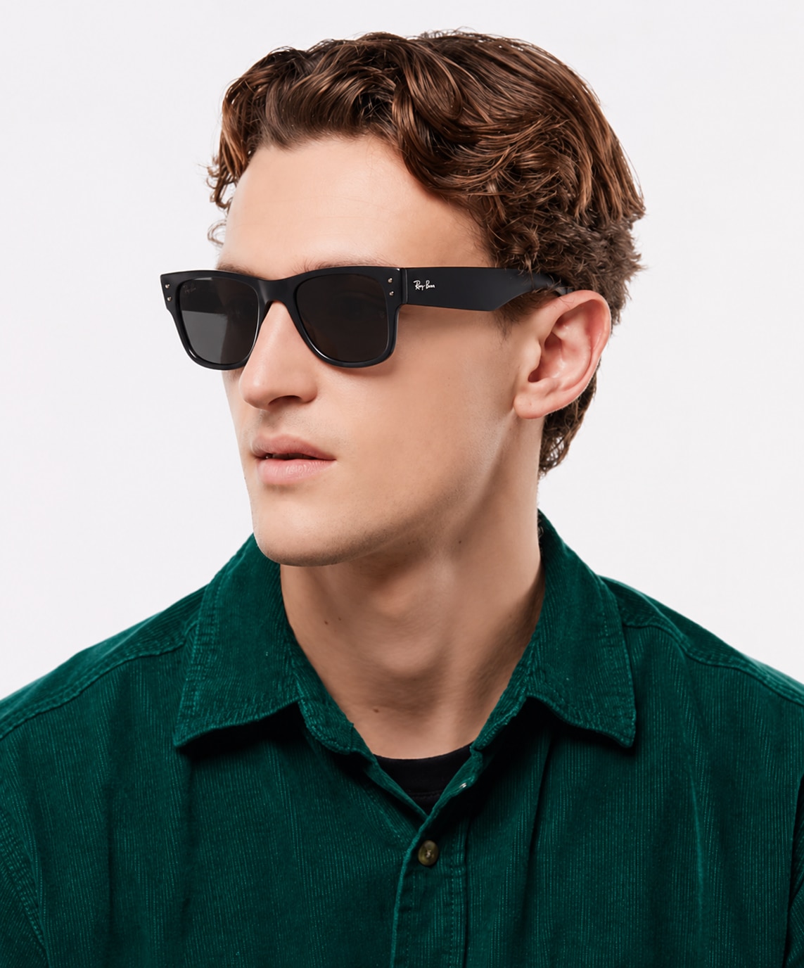 A man with short curly hair wears a dark green shirt and oversized black Wayfarer-style sunglasses, posed against a clean white studio background.