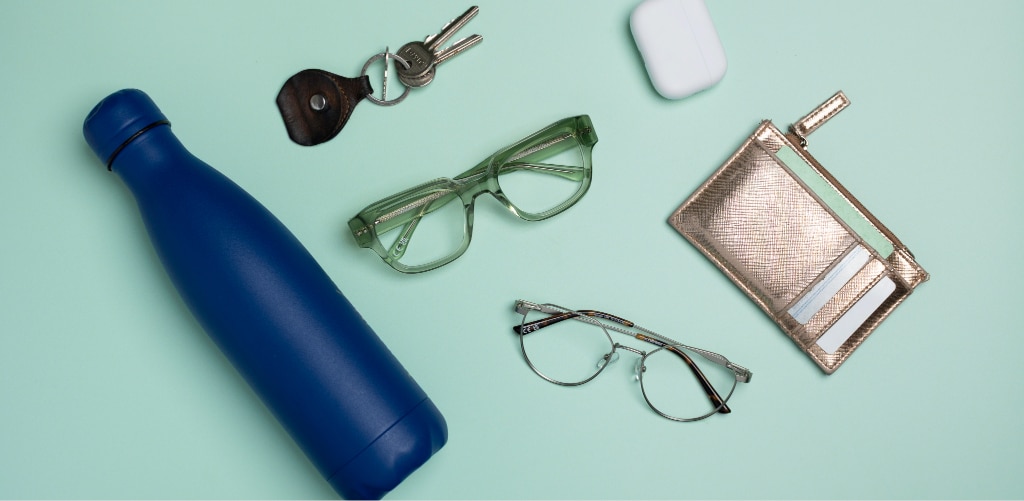 Flat lay of everyday essentials arranged on a mint green background, including a blue reusable water bottle, a set of keys, a white wireless earbud case, a gold card holder wallet, The Collection Miller transparent green square acetate eyeglasses, and The Collection Morgan of silver metal-rimmed phantos eyeglasses. The items are evenly spaced, creating a clean, organised composition.