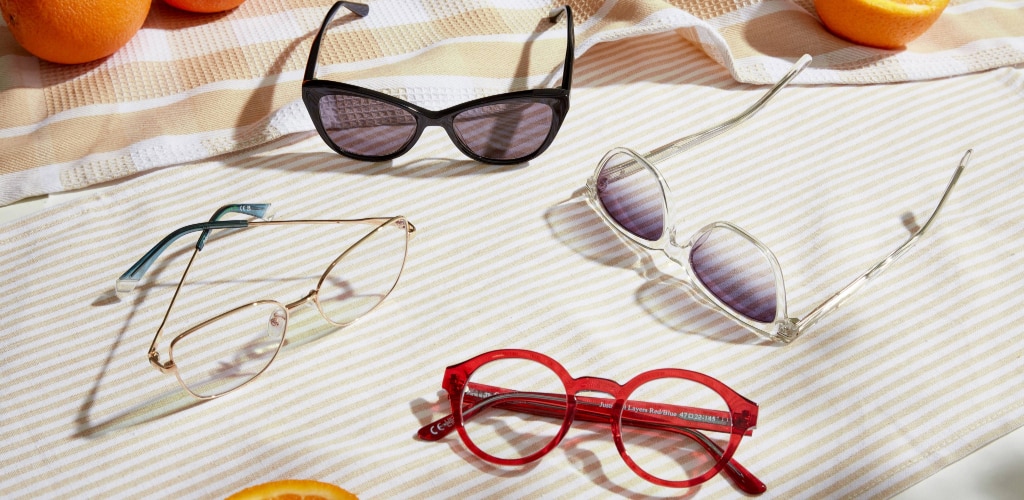 A selection of sunglasses and eyeglasses arranged on a striped cloth in bright sunlight. The styles include black cat-eye shades, gold metal cat-eye glasses, clear-framed square sunglasses, and bold red round frames. Oranges are placed around the scene, adding a fresh, summery feel.