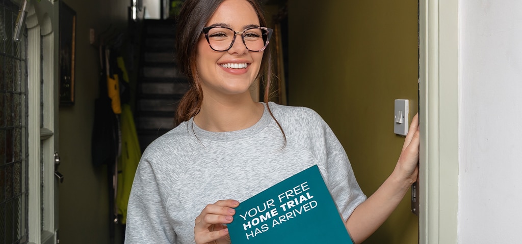 Woman with long wavy brown hair tied back, wearing large tortoiseshell cat-eye glasses and a loose grey t-shirt holding a Glasses Direct Home Trial box at her front door, smiling in natural light.