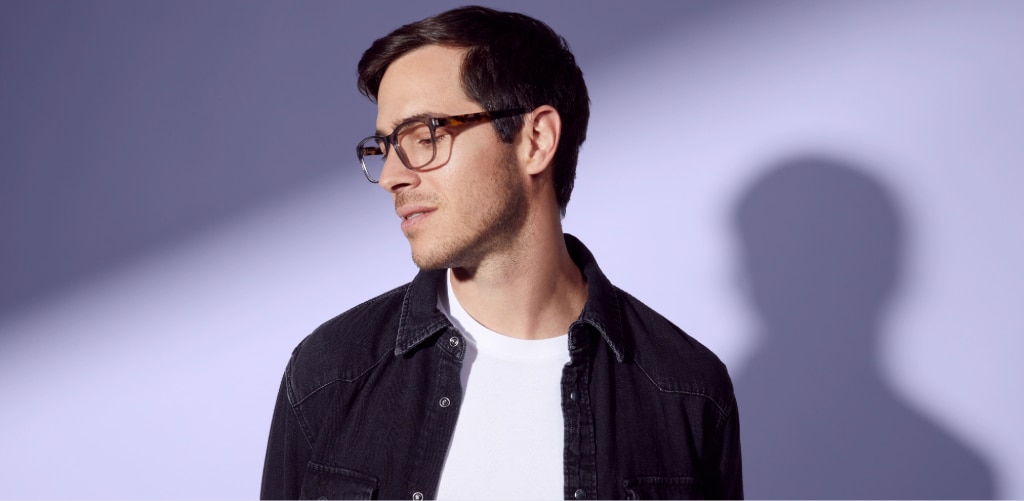 A man with short dark hair wearing tortoiseshell rectangular glasses stands against a soft lavender background with dramatic studio lighting. He's dressed in a dark denim-style jacket over a white T-shirt and is looking slightly downward and to the side. A large shadow of his profile is cast on the wall behind him, creating a stylish, fashion-focused portrait that highlights the eyewear.
