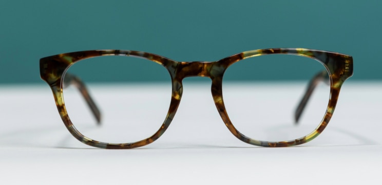 Front-facing product shot of The Collection Andi tortoiseshell glasses resting on a white surface against a teal background. The frame features a classic rectangular shape with slightly rounded edges and a distinctive keyhole bridge. The mottled brown, amber and olive-green acetate pattern gives the glasses a warm, vintage-inspired look, while the softly blurred temples in the background draw attention to the front of the frame.