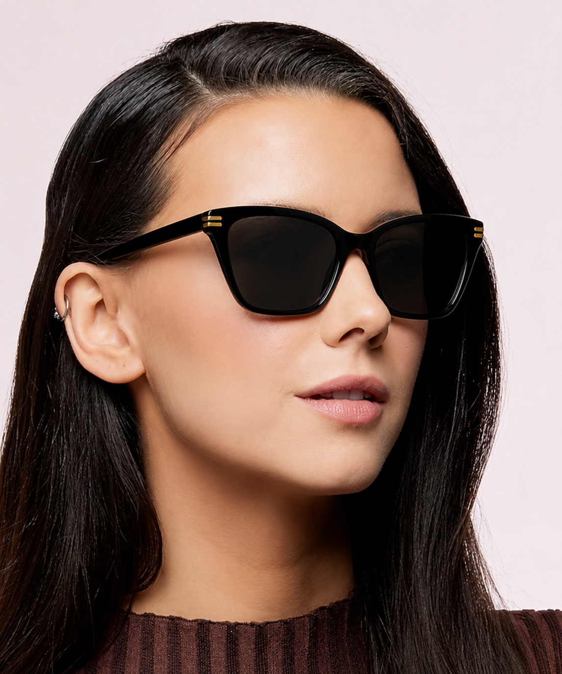 A smiling woman with medium black hair wears a brown jumper and black designer cat-eye sunglasses, posed against a clean white background.