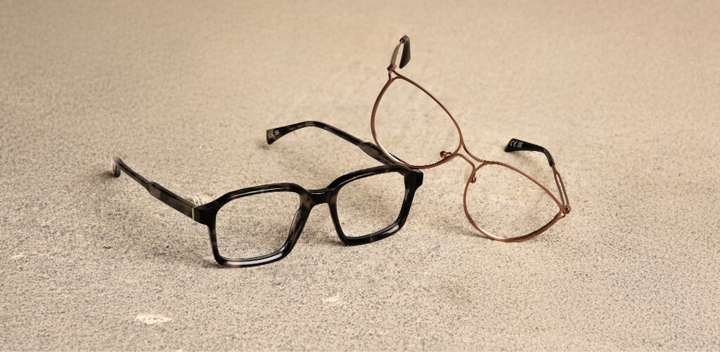 Two pairs of glasses displayed in an acetate vs metal frames comparison, featuring chunky rectangular acetate glasses alongside lightweight cat-eye metal frames. The slim metal pair leans gently against the bold acetate frames, highlighting the contrast between statement styling and minimal, refined design.