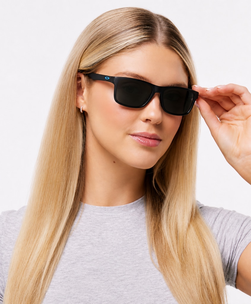 A smiling woman with long blonde hair wears a grey t-shirt and black rectangular Oakley sunglasses, posed against a clean white background.