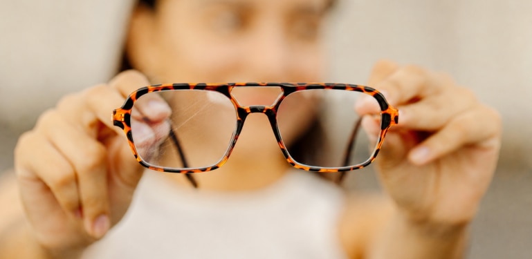 Close-up of a person holding a pair of tortoiseshell aviator glasses with visible scratches on one lens. The scratched lens appears marked and hazy compared with the clearer lens, illustrating how surface scratches can affect lens clarity and visual performance over time.