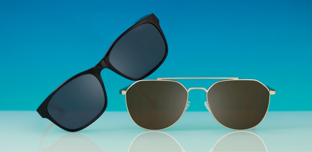 Two pairs of prescription sunglasses — black rectangular GD Collection Aero frames and gold aviator GD Colby frames — against a blue background on a reflective surface.