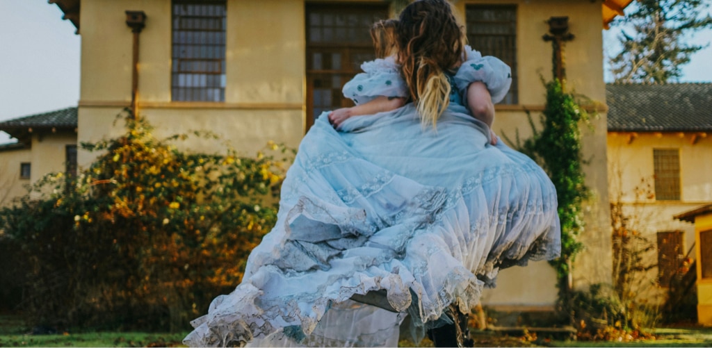 A woman with long, flowing hair is seen from behind as she runs across a garden, wearing a billowing light-blue dress with delicate lace details. Ahead stands a large, old-fashioned building with tall windows, bathed in warm afternoon sunlight. Surrounded by greenery and soft golden tones, the scene evokes the whimsical, storybook atmosphere of Alice in Wonderland.