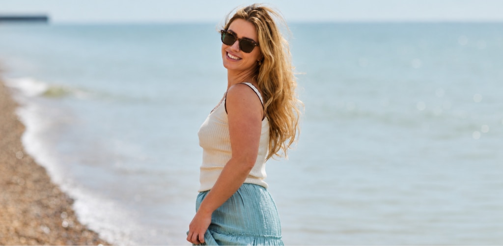 Woman with long wavy blonde hair wearing a cream vest top and light-blue maxi skirt styled with square Seaglass Sands London Retro Balham prescription sunglasses smiling on a sunny beach.