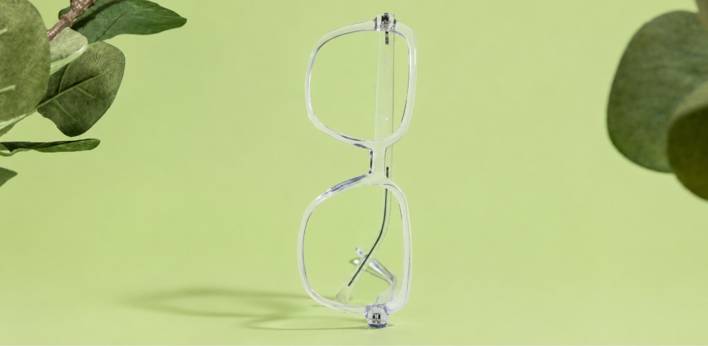 The Collection Leigh transparent bio-nylon eyeglass frames stand upright on a soft light-green surface, framed by blurred green foliage that creates a fresh, nature-inspired backdrop. The clear square-shaped frames are displayed from the side, showcasing their lightweight construction, clean lines and minimalist aesthetic. Gentle studio lighting casts a subtle shadow beneath the glasses, while the soft green tones reinforce the sustainable, eco-conscious appeal of the bio-based eyewear.