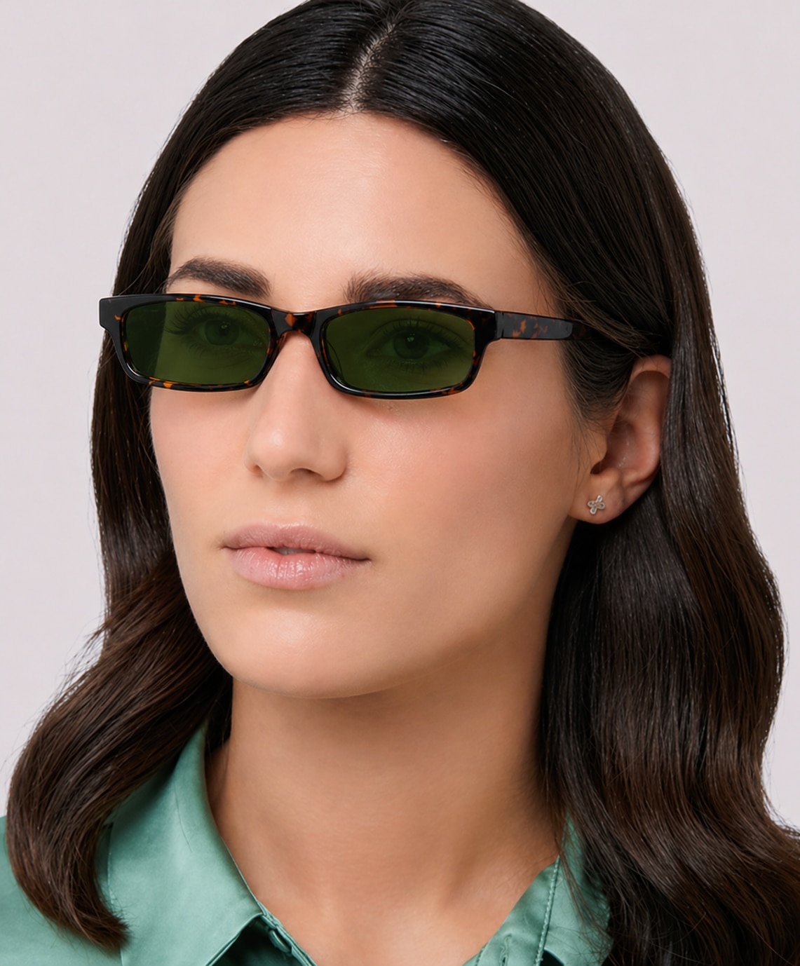 A woman with dark, shoulder-length wavy hair wears narrow tortoiseshell sunglasses, facing forward with a neutral expression against a clean, light background, dressed in a green blouse and small stud earrings.