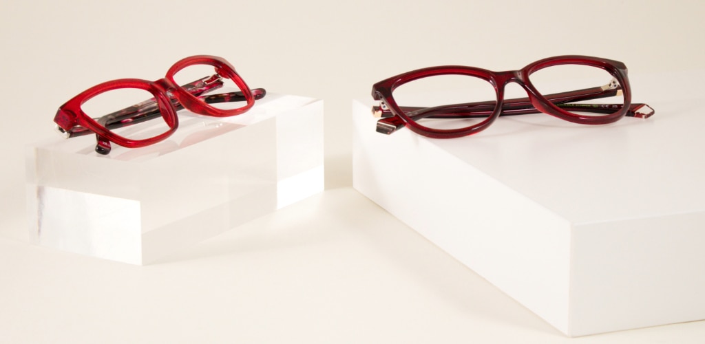 Two pairs of red and burgundy acetate glasses are displayed on white plinths against a neutral background, showcasing a bold cat-eye frame and Polaroid PLD D504 slim rectangular glasses in rich autumn-inspired colours.
