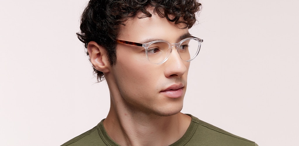 Close-up portrait of a man wearing clear acetate eyeglass frames with a rounded shape and dark tortoiseshell temples. The man has short curly dark hair and is looking to the side against a soft neutral background. The image demonstrates how clear frames offer a subtle, versatile style that complements a wide range of skin tones.