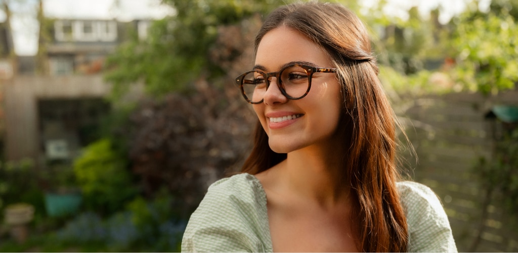 Smiling woman with long brown hair wearing round dark tortoiseshell acetate glasses and green gingham dress in a sunny garden setting.