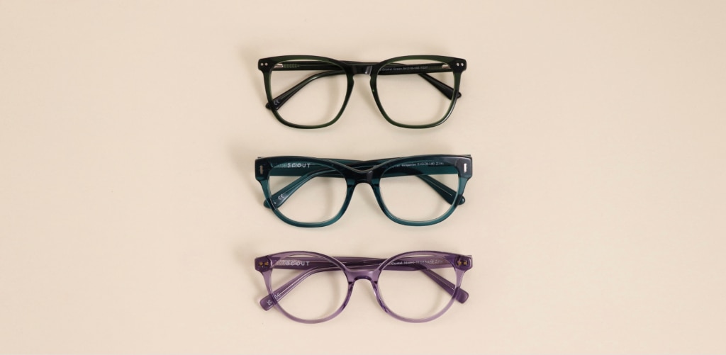 Three Glasses Direct and Scout acetate glasses frames laid flat against a warm neutral background — dark green square, teal cat-eye and transparent purple round, from top to bottom. Frames: GD Elsie, Scout Gracie and Scout Gabriella.
