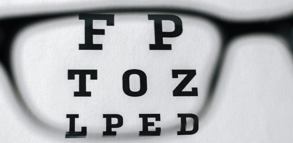 A close-up view of an eye chart seen through the lens of a pair of glasses. The letters inside the lens appear sharp and in focus, while the surrounding areas are blurred, illustrating how corrective lenses improve vision.