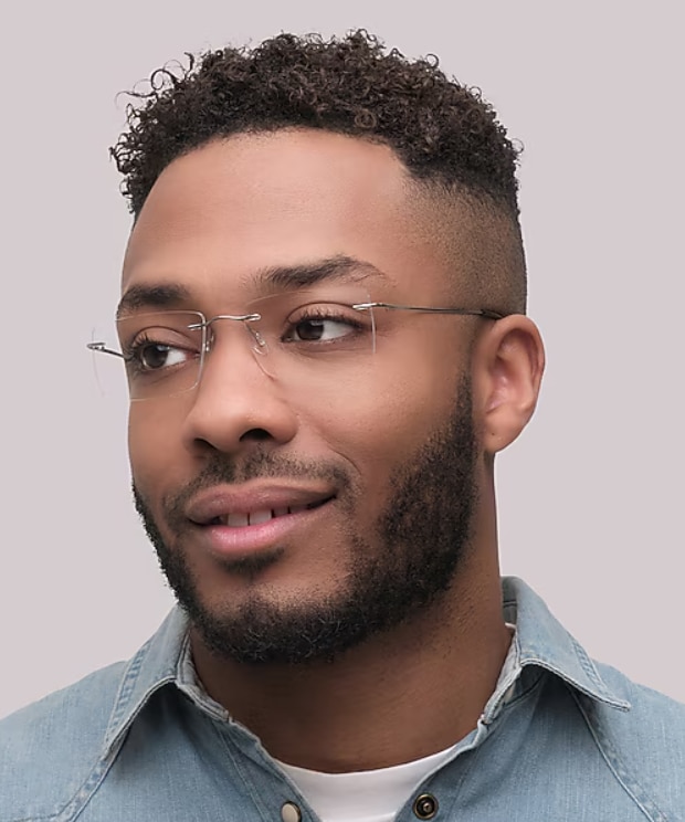 A close-up portrait of a man wearing rimless rectangular glasses with thin titanium gunmetal temples against a light neutral background. The lightweight, minimalist frame emphasises a clean and understated look. The man has short dark curly hair, a neatly trimmed beard and is dressed in a light denim shirt, creating a modern and refined appearance.
