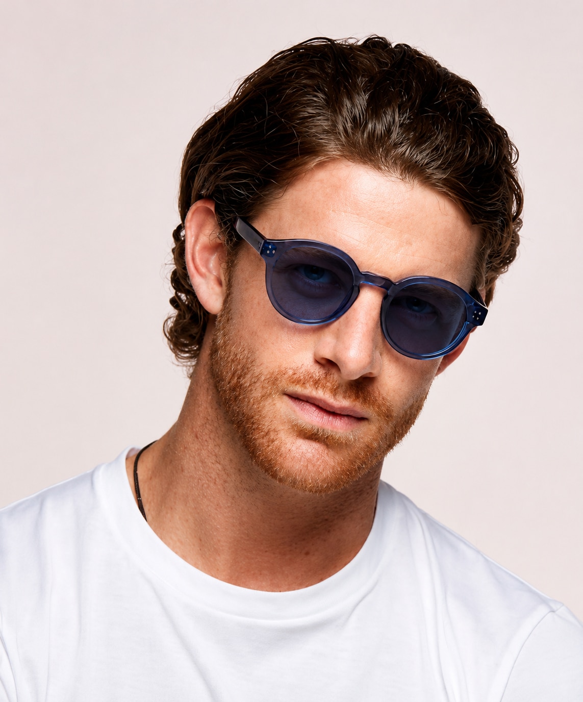 A man with medium-length wavy brown hair and light facial hair wears large round blue sunglasses with amethyst-tinted lenses and a white t-shirt. He tilts his head slightly and looks toward the camera with a relaxed expression against a plain light background.