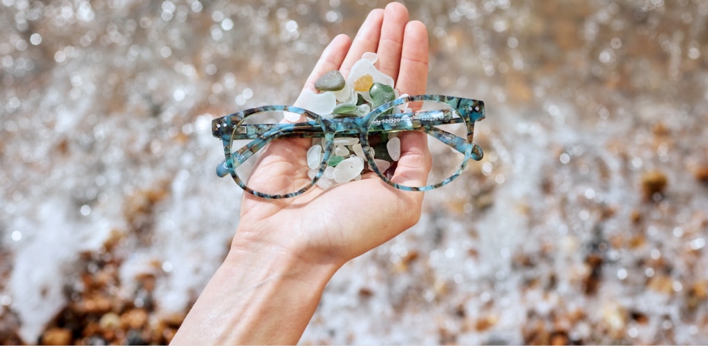 Hand holding Hart Gibson Seaglass Sands acetate phantos glasses with sea glass pebbles in front of a rocky beach background.