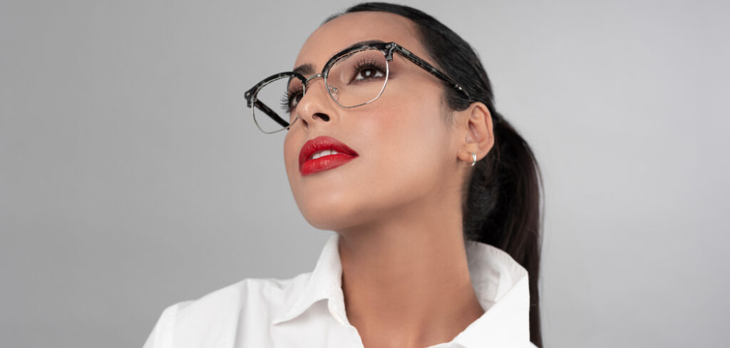 Studio portrait of a woman; wearing silver Scout Keegan square browline glasses with dark Havana grey accents along the upper frame. The person has dark hair pulled back, wears a white collared shirt and red lipstick, and is looking upward. The image is set against a light grey background with soft, even lighting that highlights the eyewear and facial features.