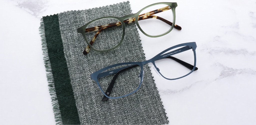 Two pairs of green glasses rest on a grey woven fabric swatch, featuring GD Whitley translucent olive-green rounded square frame with tortoiseshell temples and a matte forest-green square metal frame, showcasing rich autumn-inspired colours and finishes.