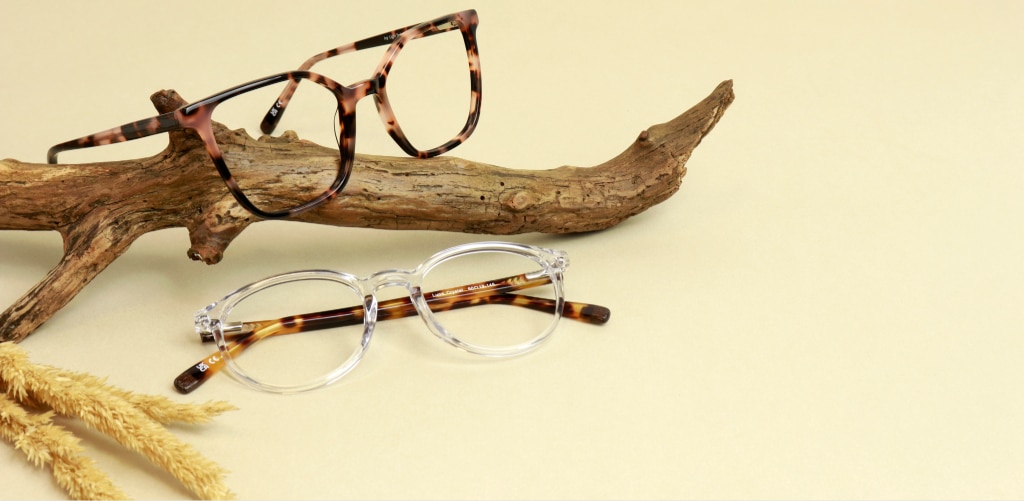 Two pairs of sustainable Arden eyeglass frames are displayed in a nature-inspired arrangement on a soft beige background. A pink tortoiseshell oversized rectangular frame rests on a piece of weathered driftwood, while a clear round frame with tortoiseshell temples is positioned below. Dried grass and natural wood elements surround the glasses, creating an organic, eco-conscious aesthetic that highlights the stylish design and environmentally friendly appeal of the eyewear.
