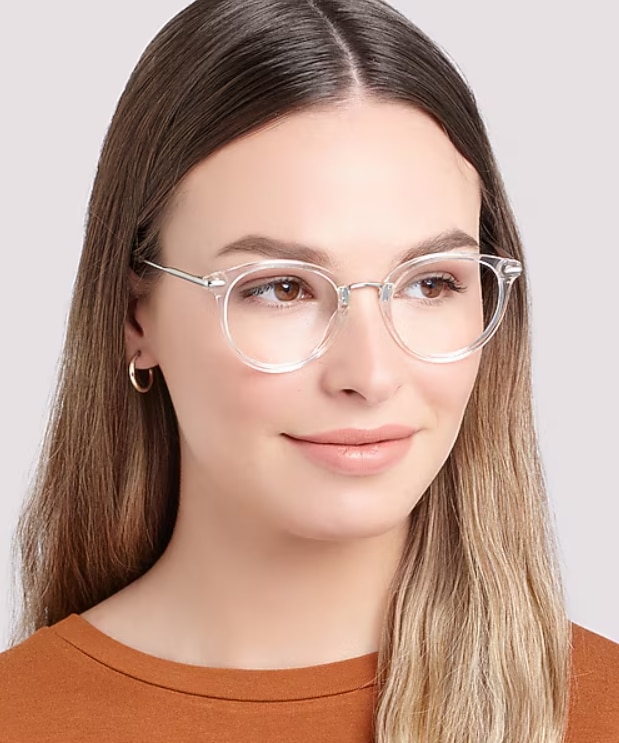 A close-up portrait of a woman wearing clear crystal round glasses with silver metal detailing against a light neutral background. The transparent frame creates a light, contemporary look that complements her natural features. With long straight hair, small hoop earrings and a warm-toned orange top, the image showcases a fresh, modern style with understated elegance.