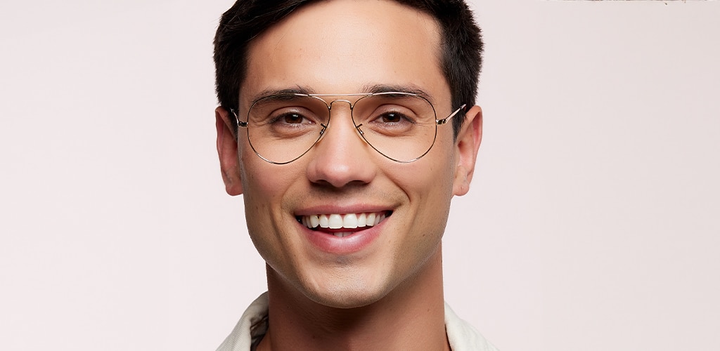 Close-up portrait of a smiling man with short brown hair wearing thin gold metal Ray-Ban® glasses with a classic aviator shape. The lightweight frame features a slim metallic finish and adjustable nose pads. Set against a soft neutral background, the image demonstrates how metal frames provide a subtle, professional appearance. 