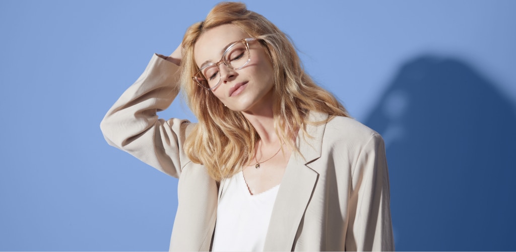 A woman with blonde wavy hair wearing clear blush-pink glasses and poses against a blue background, styled with a cream blazer and white top, showcasing transparent eyewear with a soft, contemporary look.