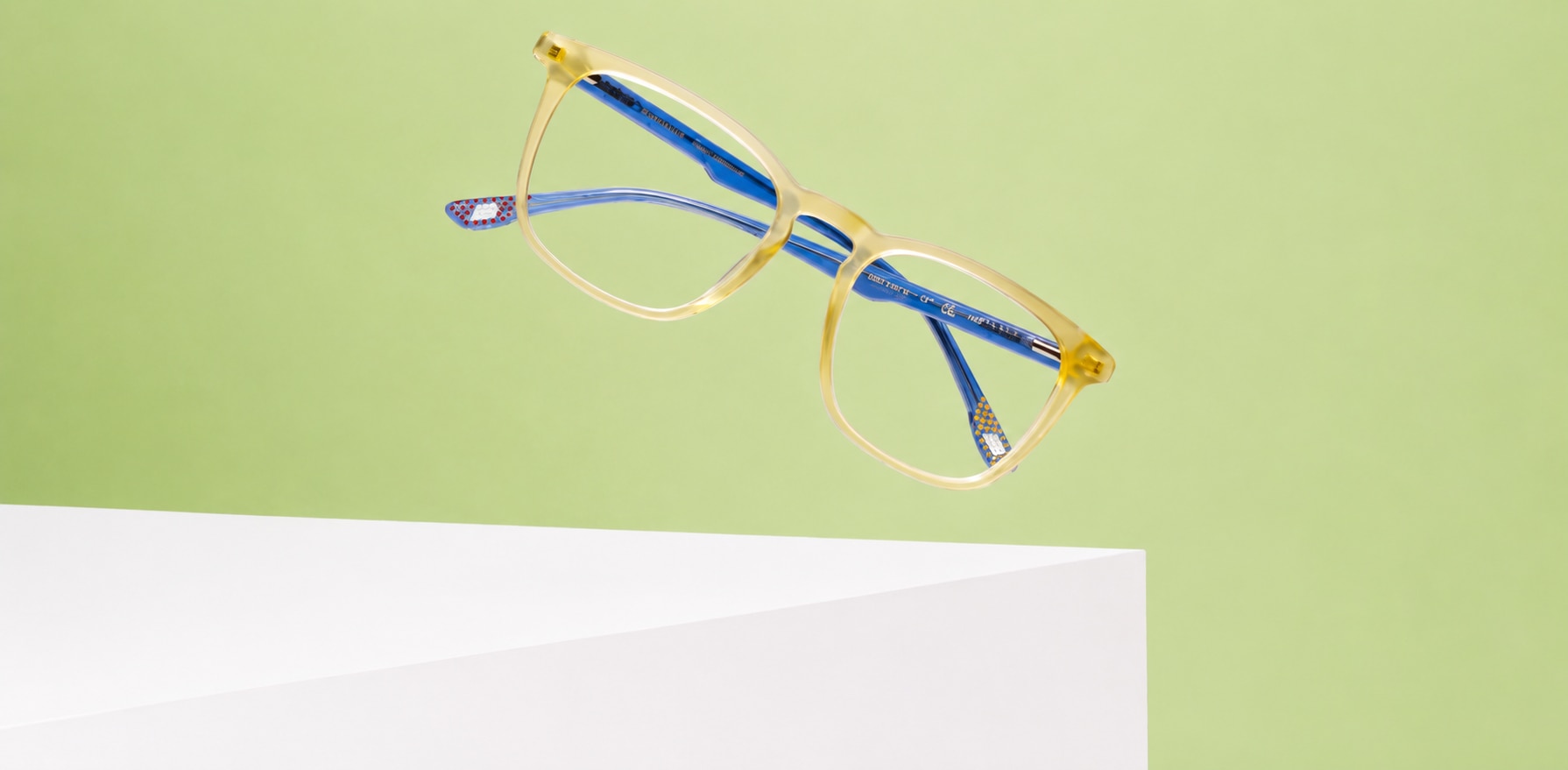 A pair of New Balance glasses with translucent yellow frames and bright blue temples appear to float above white geometric display block against a soft light-green background. The frame features a modern rectangular shape with gently rounded edges, while the vibrant blue temple arms add a playful contrast. Styled as a contemporary product photograph, the image uses clean lines, soft lighting and a fresh colour palette to highlight the eyewear's lightweight design and distinctive details.