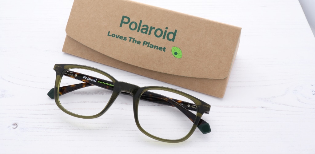A pair of olive-green Polaroid sustainable eyeglass frames is displayed on a white wooden surface beside a kraft-paper glasses case printed with the message “Polaroid Loves The Planet.” The glasses feature a rounded-square shape with translucent green rims and tortoiseshell-patterned inner temples. The words “Sustainable Choice” are visible on the inside of the frame arm, reinforcing the eco-friendly theme. The natural cardboard case, green branding and clean, minimalist styling highlight the environmentally conscious design and sustainable materials of the eyewear.