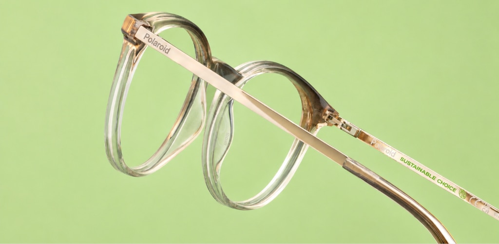 Close-up of a pair of sustainable Polaroid eyeglass frames against a soft light-green background. The translucent frame features warm amber-brown accents and slim gold-tone metal temples, with the words “Sustainable Choice” visible on the arm. The glasses are photographed at an angle, highlighting the rounded lens shape, lightweight construction and eco-conscious design. Soft lighting and the fresh green backdrop reinforce the environmentally friendly aesthetic while showcasing the frame's modern, refined style.