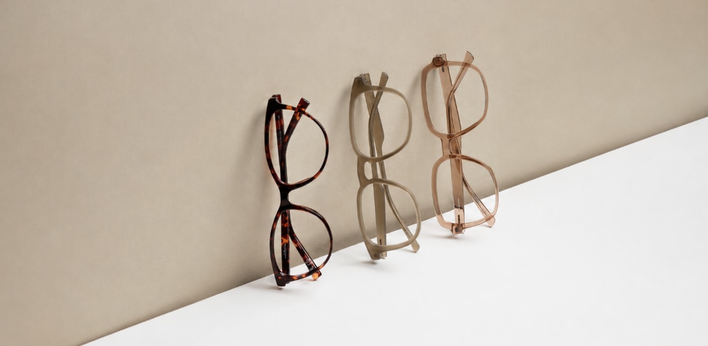 Three pairs of Glasses Direct frames are displayed upright against a soft beige backdrop. The frames feature different finishes—a Havana pattern, a muted matte green and a translucent shiny beige—arranged on a clean white surface. The minimalist composition and neutral tones create a refined, contemporary luxury aesthetic.