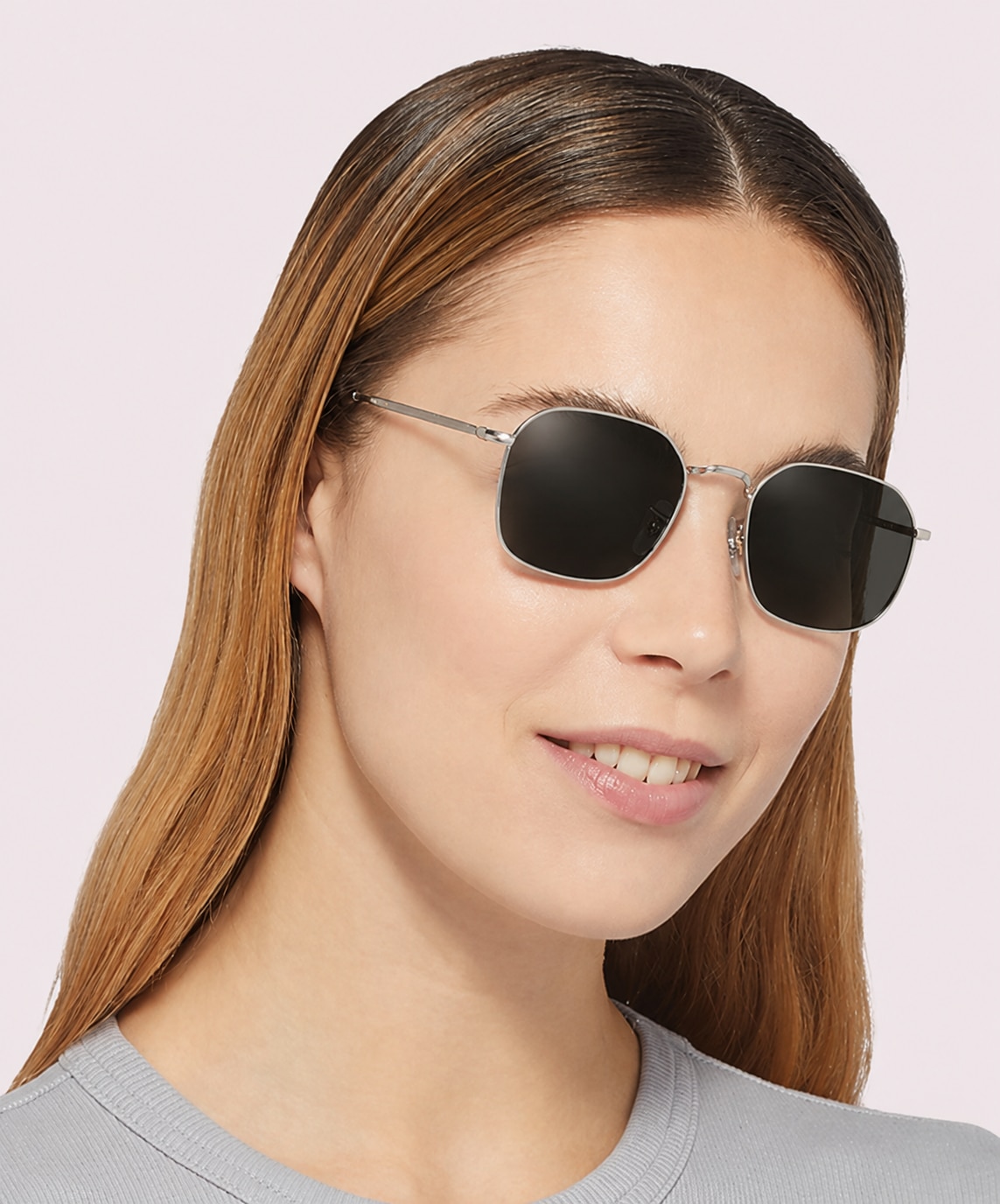 A smiling woman with long blonde hair wears geometric silver metal sunglasses, dressed in a grey top, against a clean, light background.